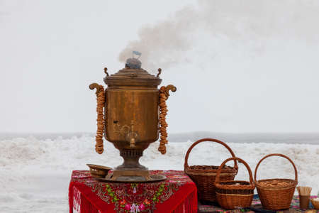 A smoking antique samovar stands on a table covered with a red tablecloth.