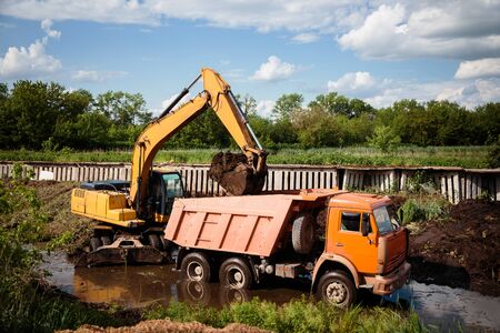 Large excavator loads earth, soil into the back of truck using bucket. Construction equipment in the water. Construction work, creating the landscape of the locality.の写真素材