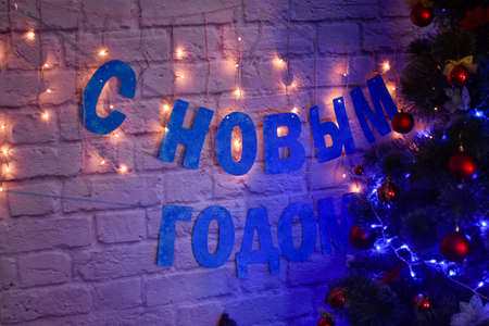 Happy new year in Russian on a brick background next to a Christmas tree lit with a garlandの写真素材