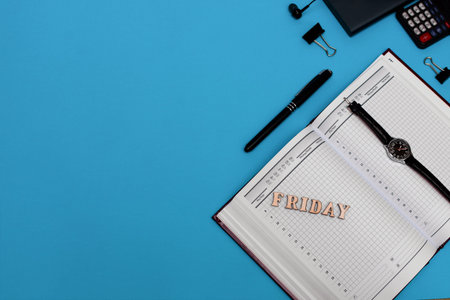 Blue work desk with blank notebook and office supplies. Top view with copy space. Friday lettering.