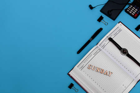 Modern office desk workplace with blank notebook, pen and supplies. Copy space on blue background. Top view. Flat lay style. Inscription sundayの写真素材