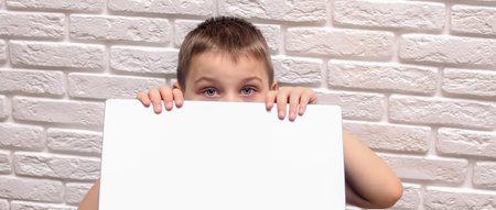 The boy looks out from behind his laptop. On the background of a white brick wall.の写真素材