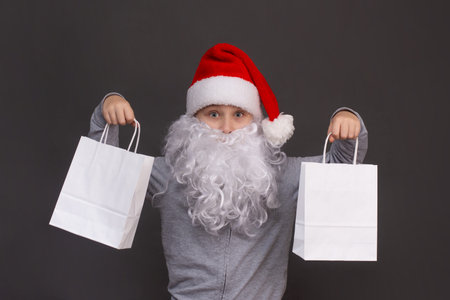 little Santa boy is holding two white paper bags in his hands. on a gray background.