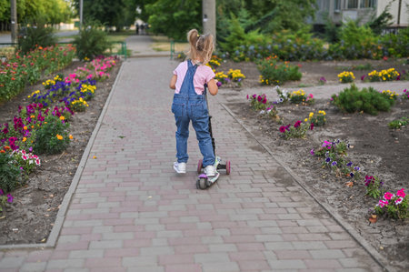 A vertical photo of a little girl riding a scooter on a street path. The image captures her joyful and carefree movement, highlighting the simplicity and happiness of childhood outdoor play.の写真素材