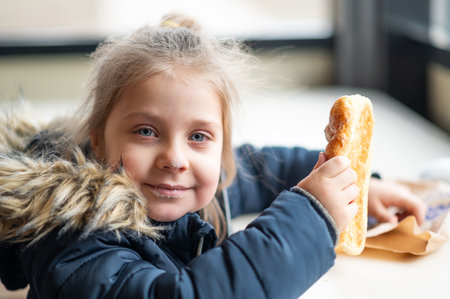 little girl sits at a table in a cafe and eats a delicious crunchy bun. delicious food for a child. a girl in a cafeteria in a winter jacket.の写真素材