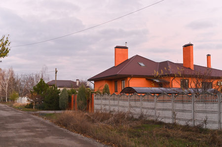 Country house behind long fence near empty road, house roof glowing in sunset light, rural residential scene. outdoor lifestyle concept, autumn season. Private country house.の写真素材