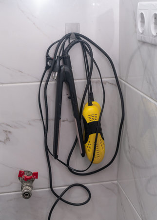Water valve with red handle, hair straightener in the bathroom, mounted on tiled bathroom wall beside black cable. verticalの写真素材