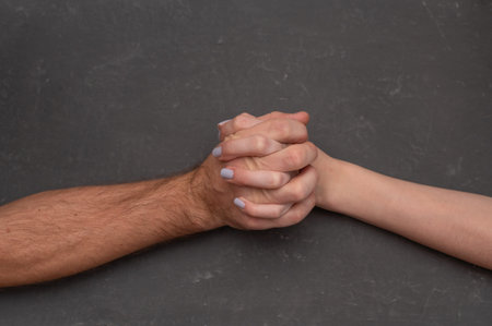 Clasped hands of two people gripping each other over dark background, hands showing support and...