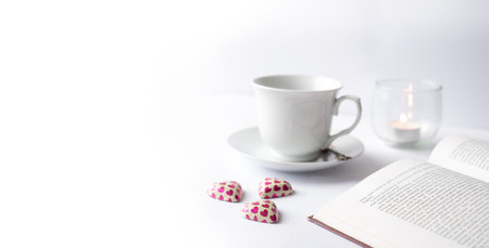 Beautiful white relaxing reading scene with open book, cup, candle and heart shaped chocolate candy.の写真素材
