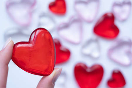 Fingers holding a red heart with blurred hearts in background. Valentine`s day background. Shiny glass hearts on white background.の写真素材