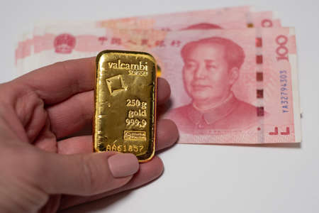 Hand holding 250 gram gold bullion bar with Chinese Yuan renminbi banknotes in the background. Money and currency. Bank of China reserves. Gold backed Chinese yuan. Financial system of China.の写真素材