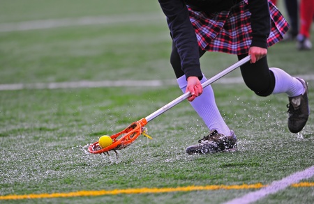 High school girls varsity lacrosse player scoops up the ball on a rain drenched turf field.の写真素材