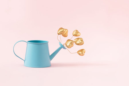 Golden hearts blooming out of blue flower watering can against pastel pink background. Creative minimal love concept.