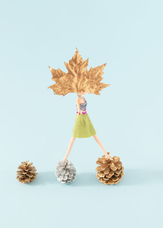 Creative composition made with three pinecones and a doll holding golden maple leaf against a bright...