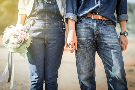 Young loving couple holding hands each other with wedding bouquet flowers , Bride and groom in vintage jeans standing together in soft light of sunsetの写真素材