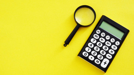 Calculator with magnifying glass for accountant on yellow background. Business and education concept. Empty space for text and logoの写真素材
