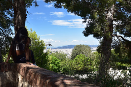 A young woman is sitting on a stone wall and looking at the city of Barcelonaの写真素材