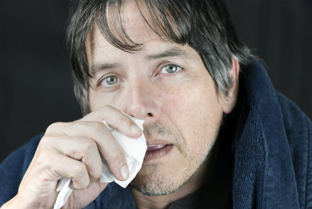 Close-up of a sick man in a housecoat dabbing his runny nose with a tissue.