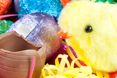 Close-up of a yellow fluffy chick toy beside a chocolate egg with a pile of colorful eggs and...