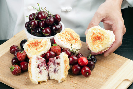 Close-up of a chef displaying a cream cheese iced strawberry cherry muffin surrounded by fruit and...