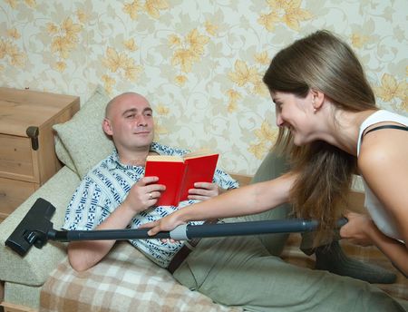 wife is cleaning up whan her husband reading a book over sofaの写真素材