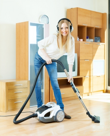 Happy blonde woman in headphones cleaning with vacuum cleaner on parquet floor at homeの写真素材
