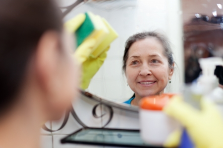 mature woman cleans mirror with sponge in bathroom at home. Focus on reflectionの写真素材