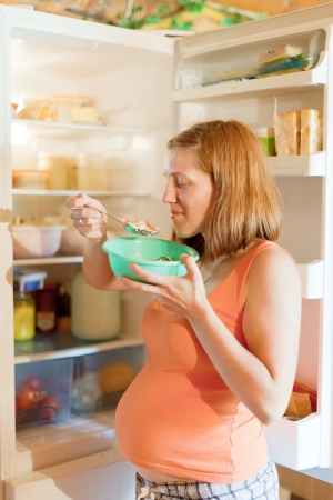 pregnant woman eating  from dish near  refrigerator at homeの写真素材