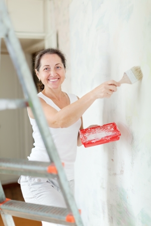 Happy mature woman paints wall with brush at homeの写真素材