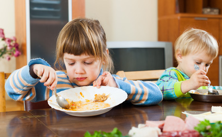 Two serious  children eating food at wooden tableの写真素材