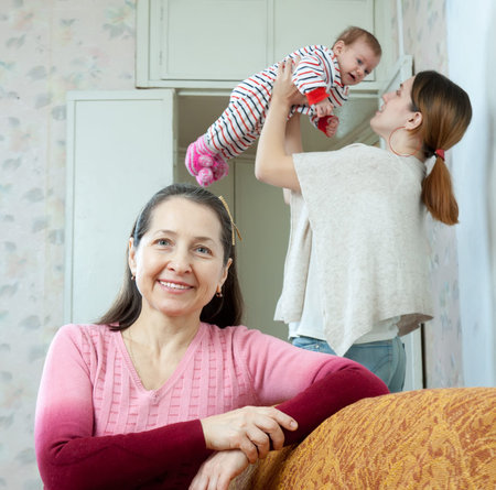 Portrait of happy mature woman against her adult daughter holds little baby at home interior
