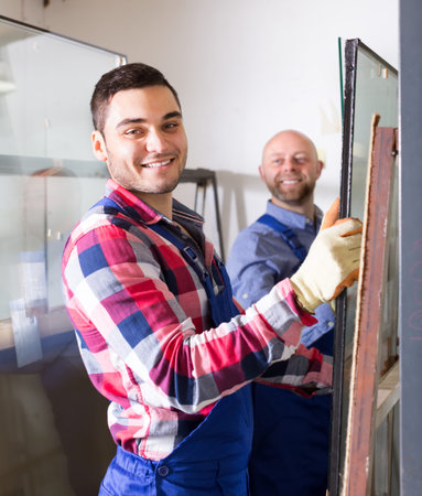 Two positive smiling workmen in coverall working with glass at factory
