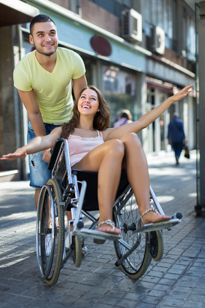 Husband taking happy young spouse on wheelchair in playful mood outsideの写真素材