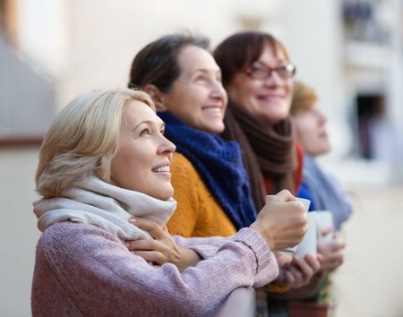 Smiling elderly woman in warm clothes having cup of hot tea on terrace. Focus on blonde woman