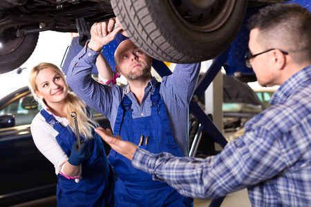 Driver has brought his car to a workshop. Mechanics a man and a woman are repairing suspension on a lifted up carの写真素材