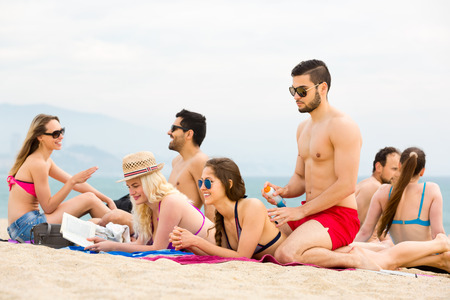 Group of people resting on a beach on a shore of a sea. Man is rubbing a sunscreen into the back of a womanの写真素材