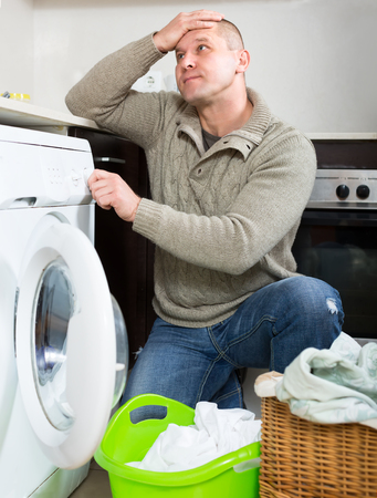 Man is holding his head sitting near washing machine thinking of how to use it. Guy doing laundryの写真素材