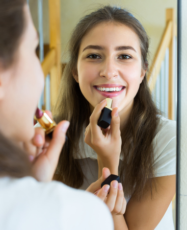 Happy young teenager applying lipstick in front of a mirror at homeの写真素材