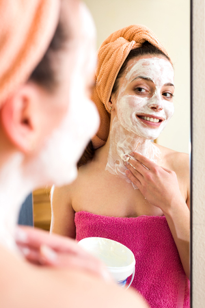 Portrait of a smiling woman in a bathroom in a mirror wrapped in a pink towel putting a skin care mask on her face and neckの写真素材