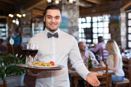Smiling young male waiter serving restaurant guests at tableの写真素材