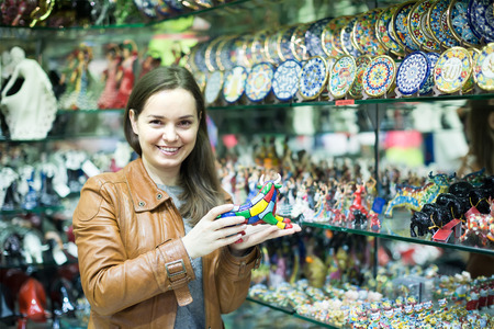 Cheerful young woman in leather jacket choosing things for memory in souvenir shopの写真素材