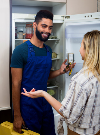 Young housewife and black handyman standing near fridge in flatの写真素材