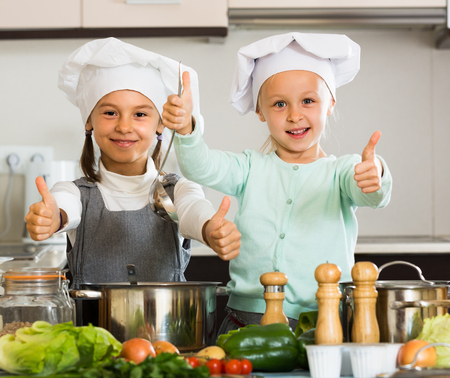 Happy little sisters in caps learning how to cook at home kitchenの写真素材
