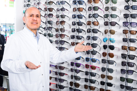 Mature male ophthalmologist standing near display with glasses in optics store