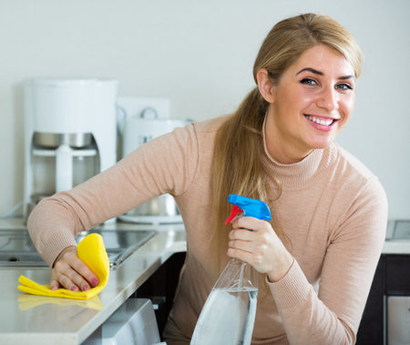 Joyful blonde maid cleaning in domestic kitchen with sprayer and smiling