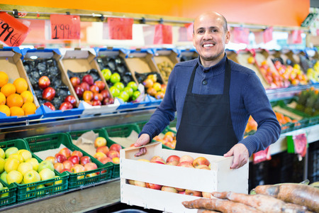Elderly friendly smiling  seller offering seasonal ripe fruits in local groceryの写真素材