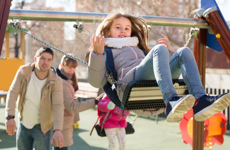 Happy young parents watching smiling little daughters swinging at playgroundの写真素材