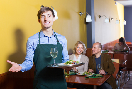 Professional happy waiter with tray posing at table of senior customersの写真素材
