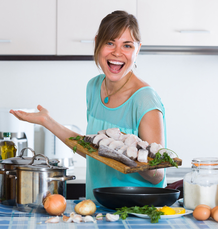 Cheerful young housewife at kitchen putting fish in frying panの写真素材