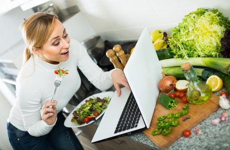 Portrait of busy young woman eating salad with laptop in kitchenの写真素材
